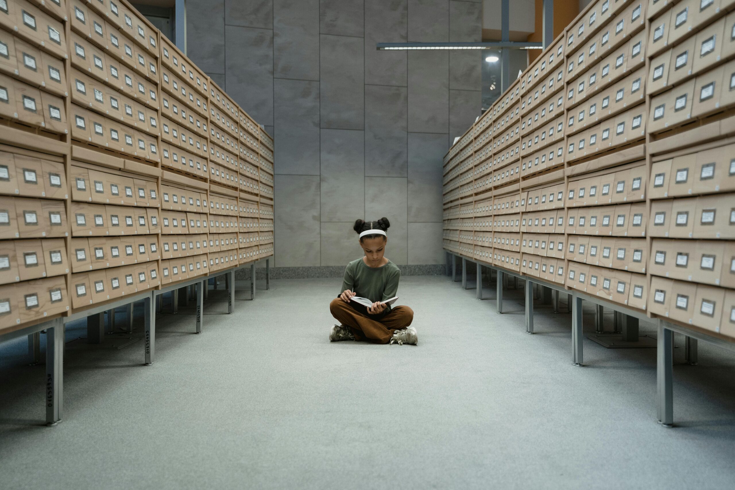 A girl sits reading a book in a library archive with wooden drawers.