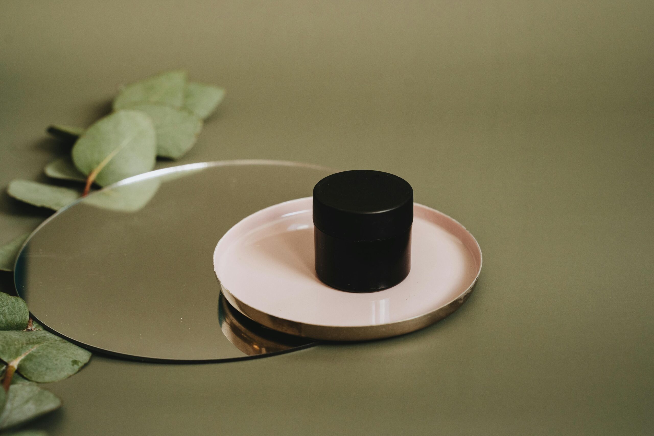 A minimalist display of a skincare product on a tray with eucalyptus leaves on a reflective surface.