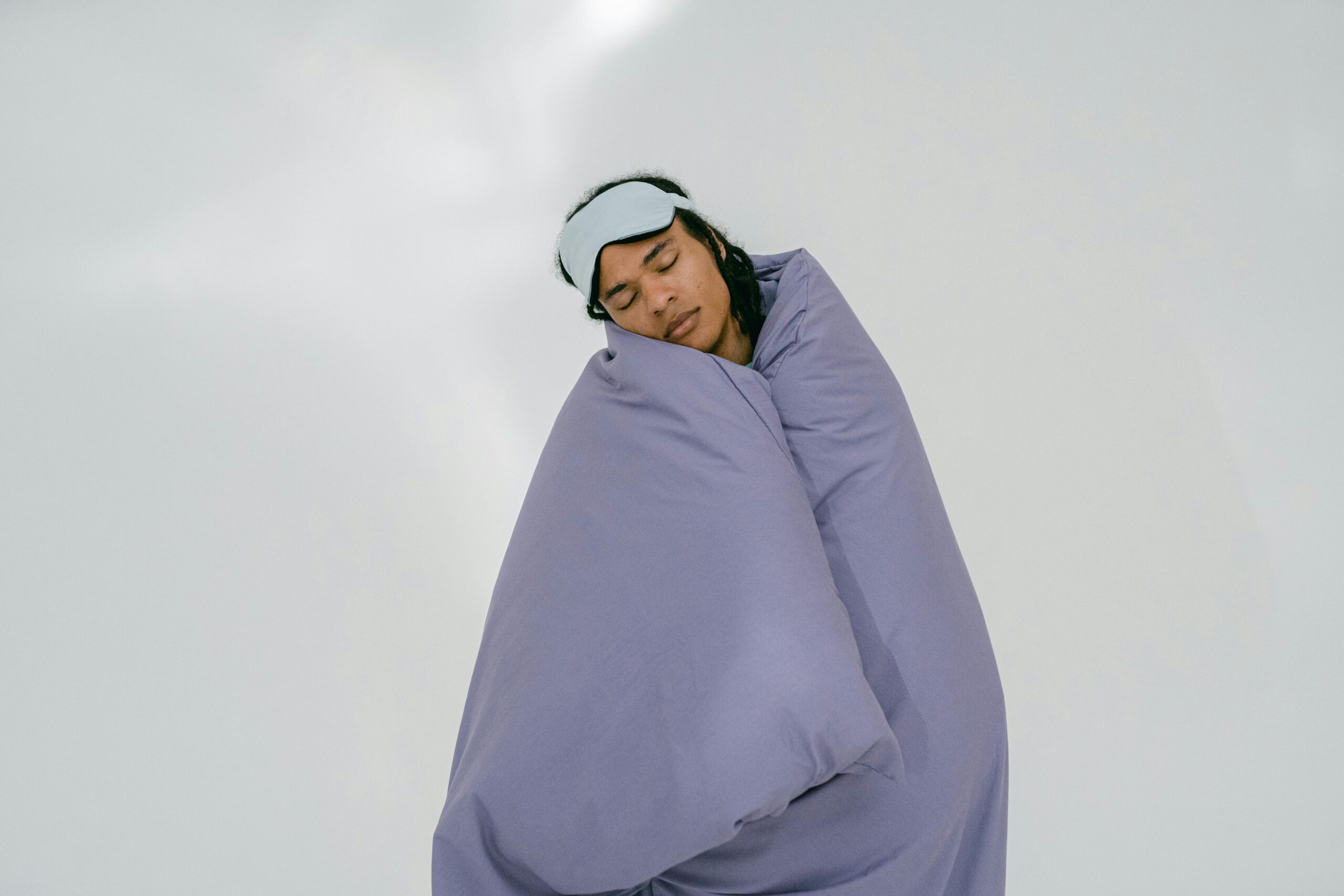 A person peacefully sleeping with an eye mask, wrapped in a purple blanket on a white background.