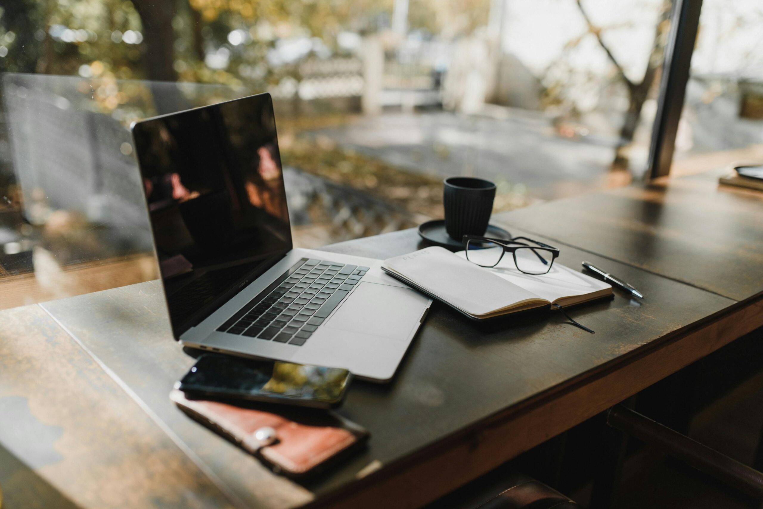 A serene office setup with a laptop, notebook, and coffee near a window, reflecting an autumn scene.