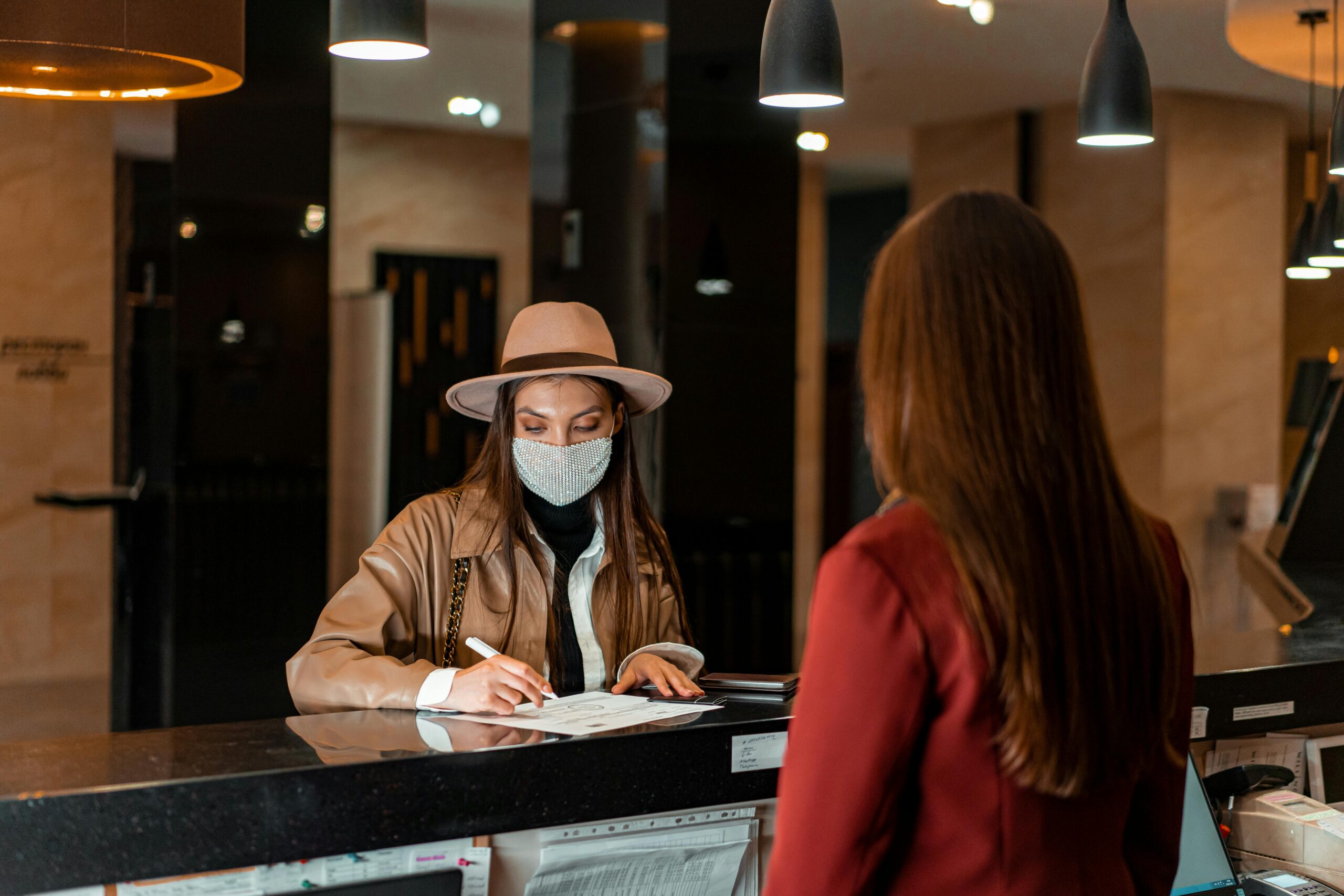 A woman in a hat and face mask checks in at a hotel reception, interacting with a receptionist. Регистрация гостей и согласие на обработку персональных данных.