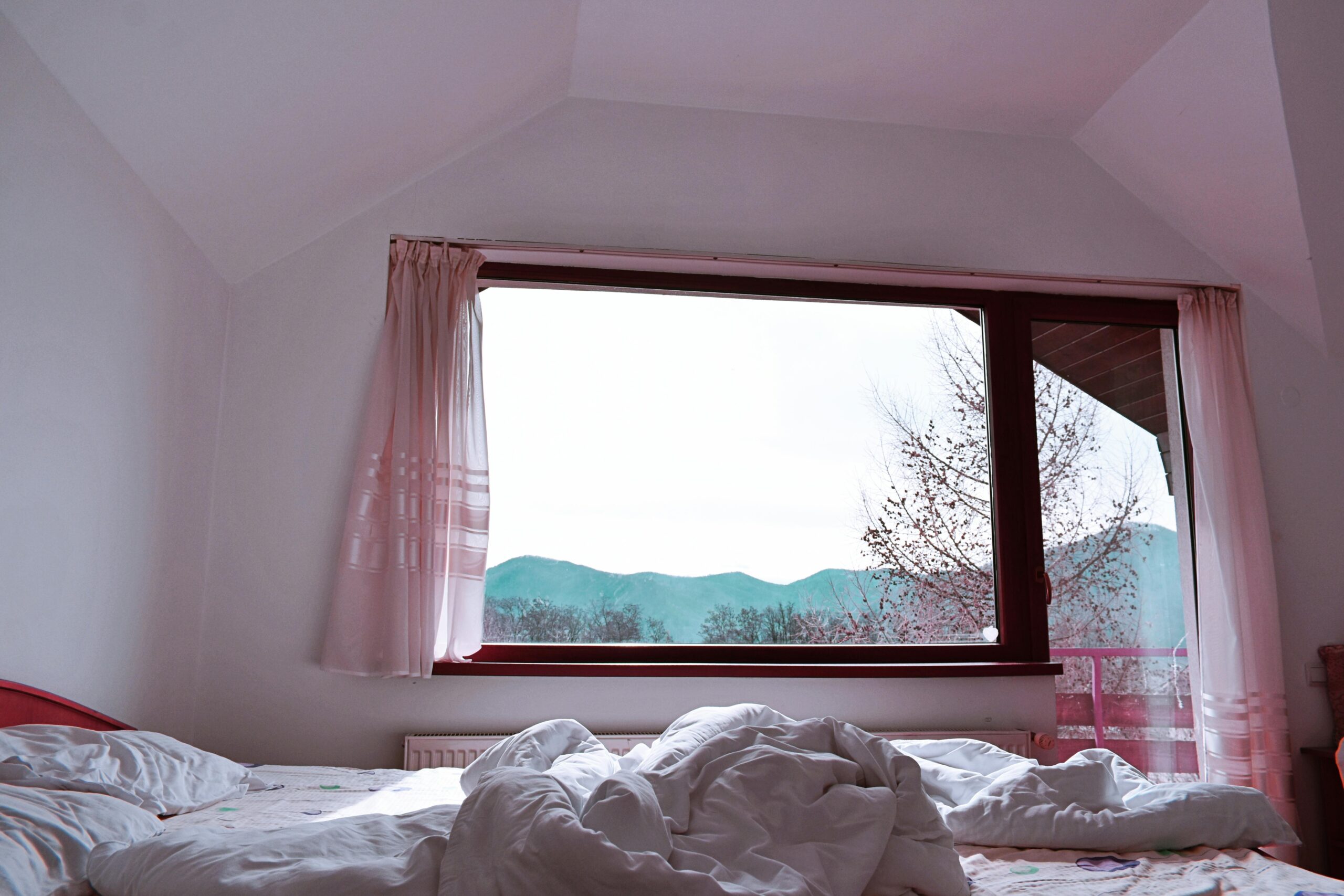 Bright bedroom interior with rumpled bed facing mountain view from window.
