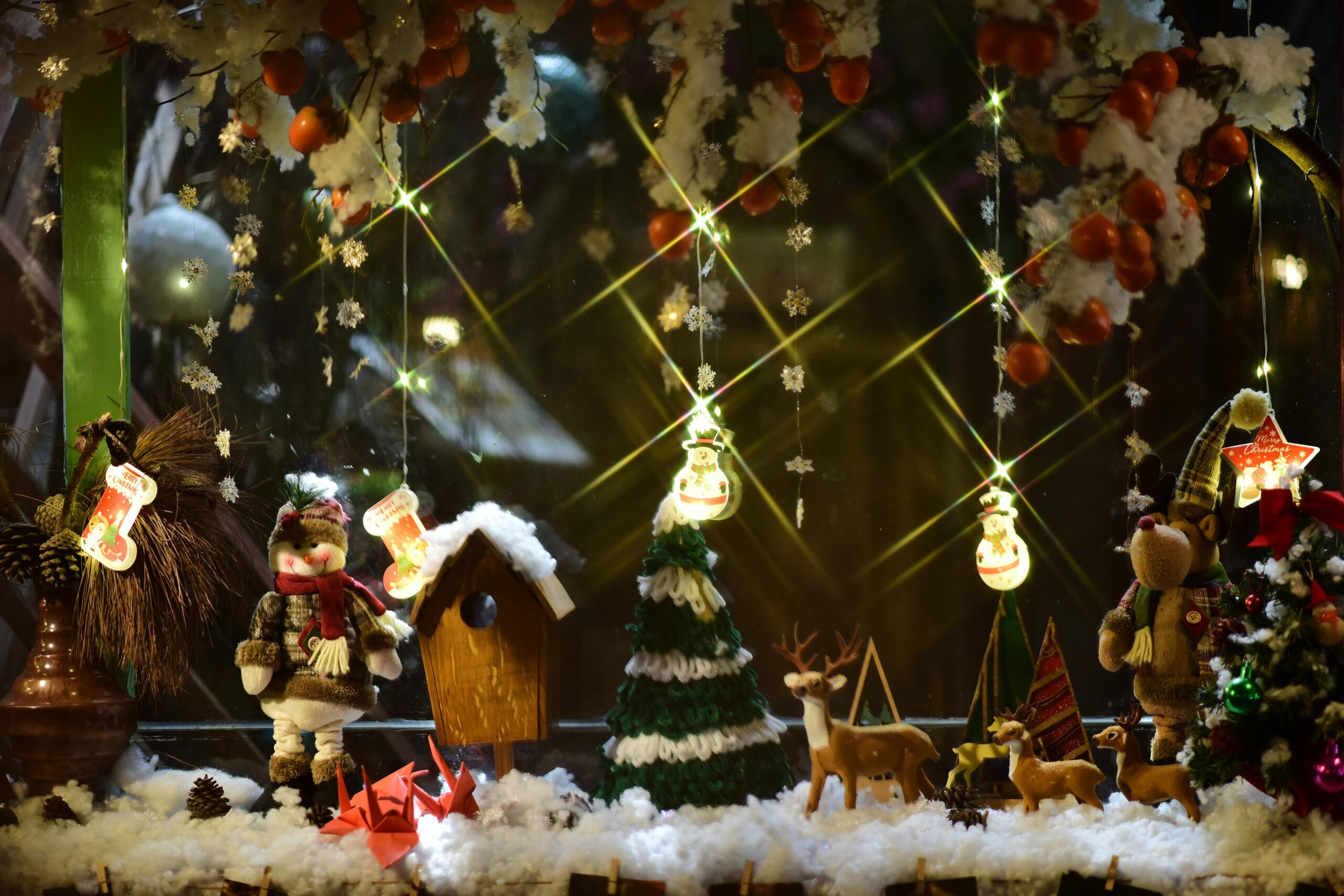 Charming holiday window display featuring festive figurines, lights, and decorations in winter setting.