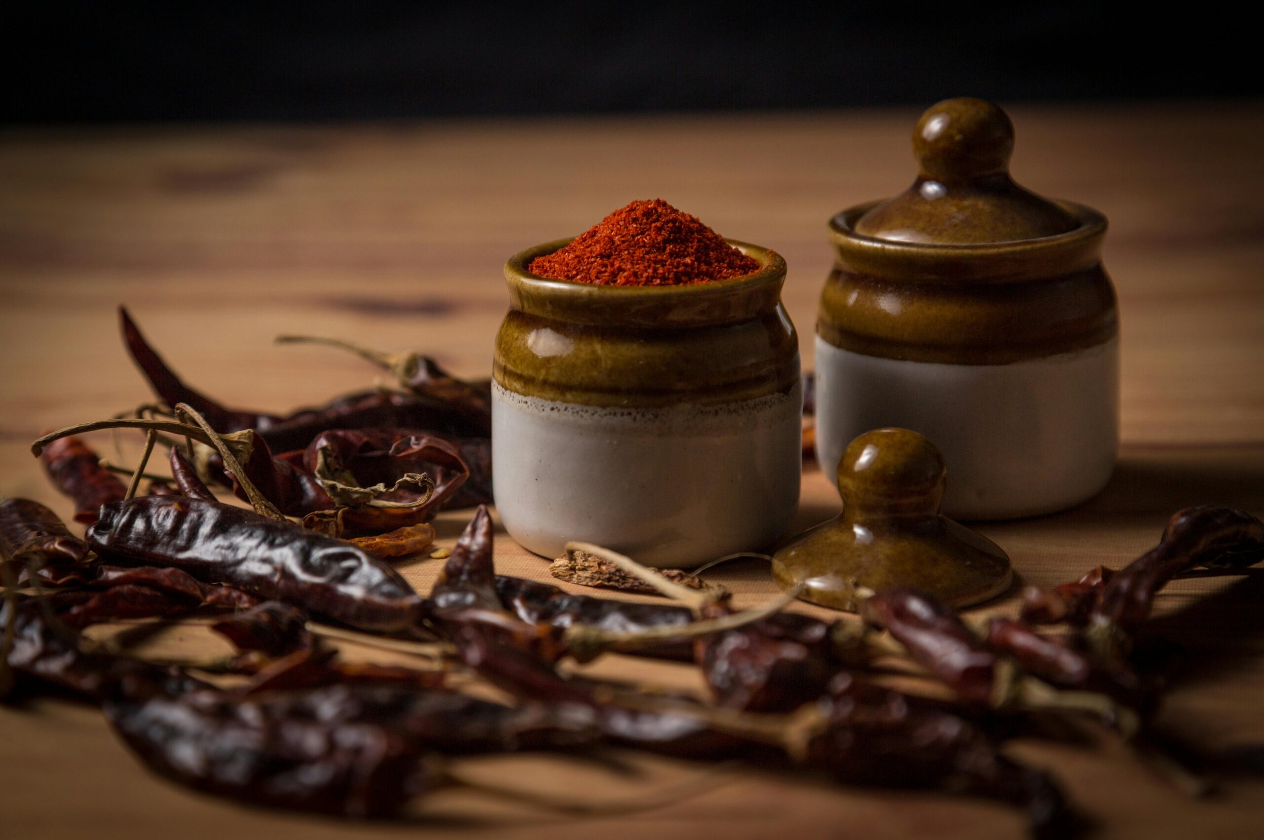 Close-up of rustic spice jars with paprika surrounded by dried peppers on a wooden surface.