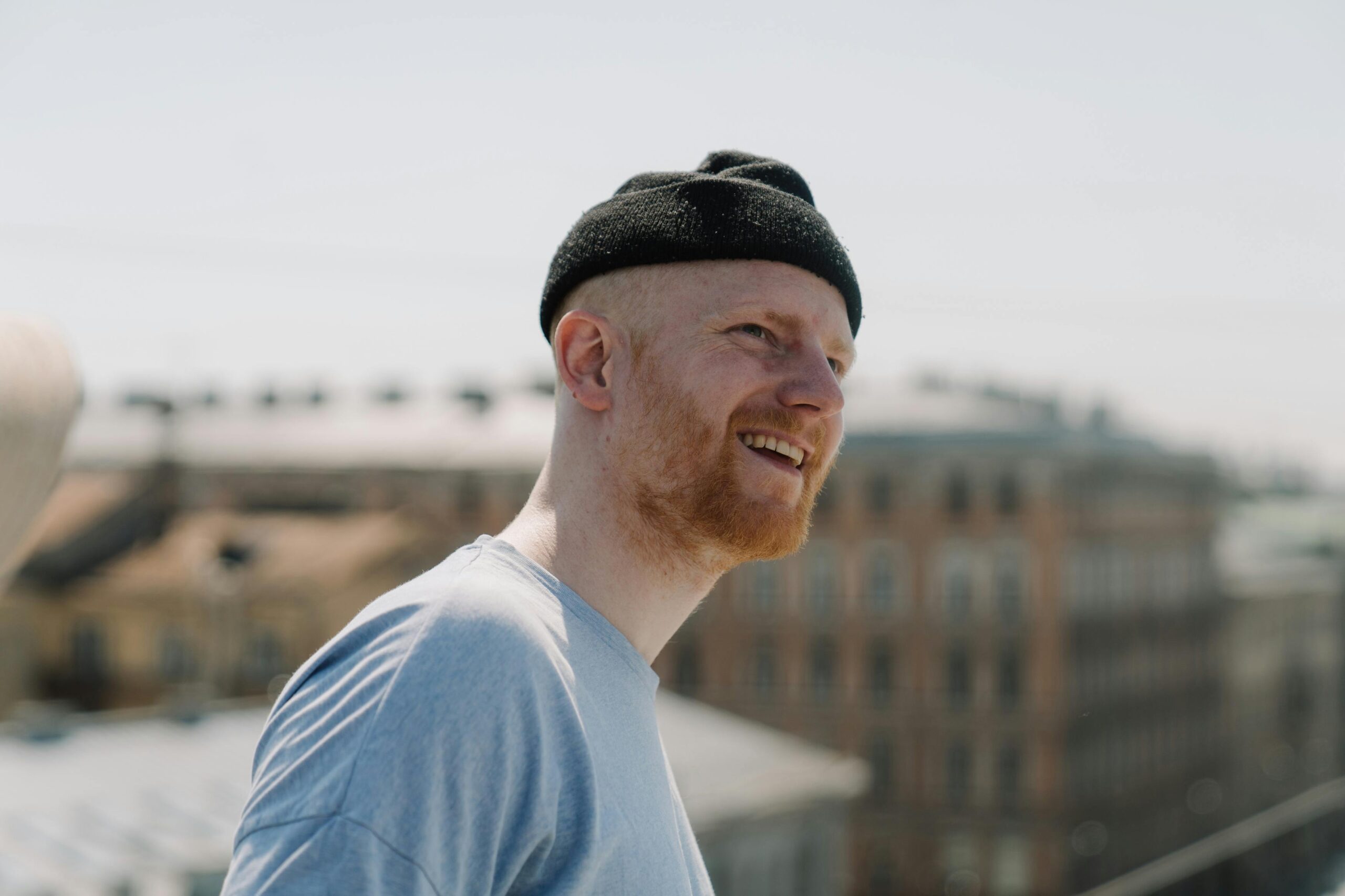 Happy man with beard and beanie smiling on a rooftop with city background.