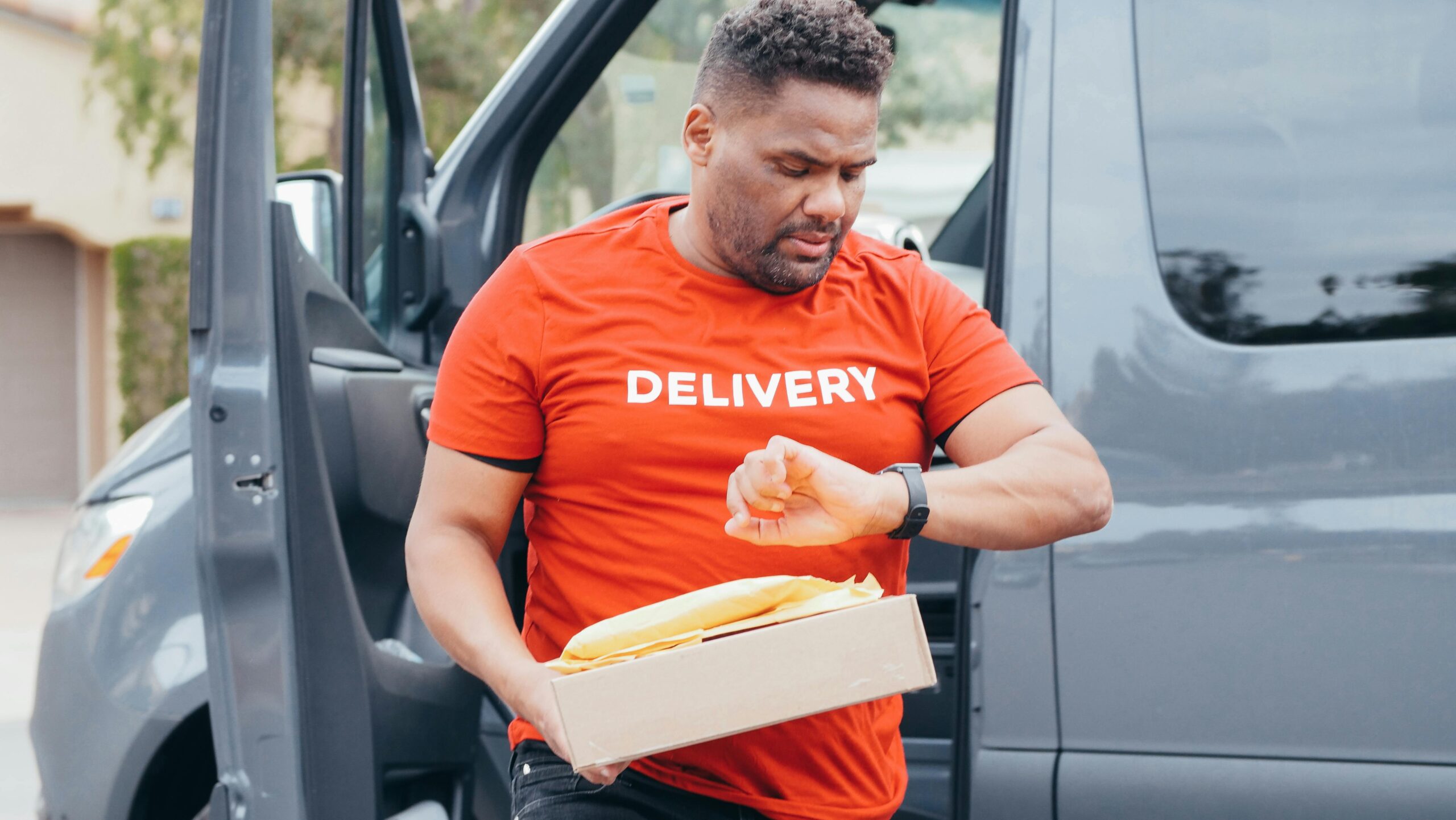 Delivery driver checks watch while carrying package from van outdoors.
