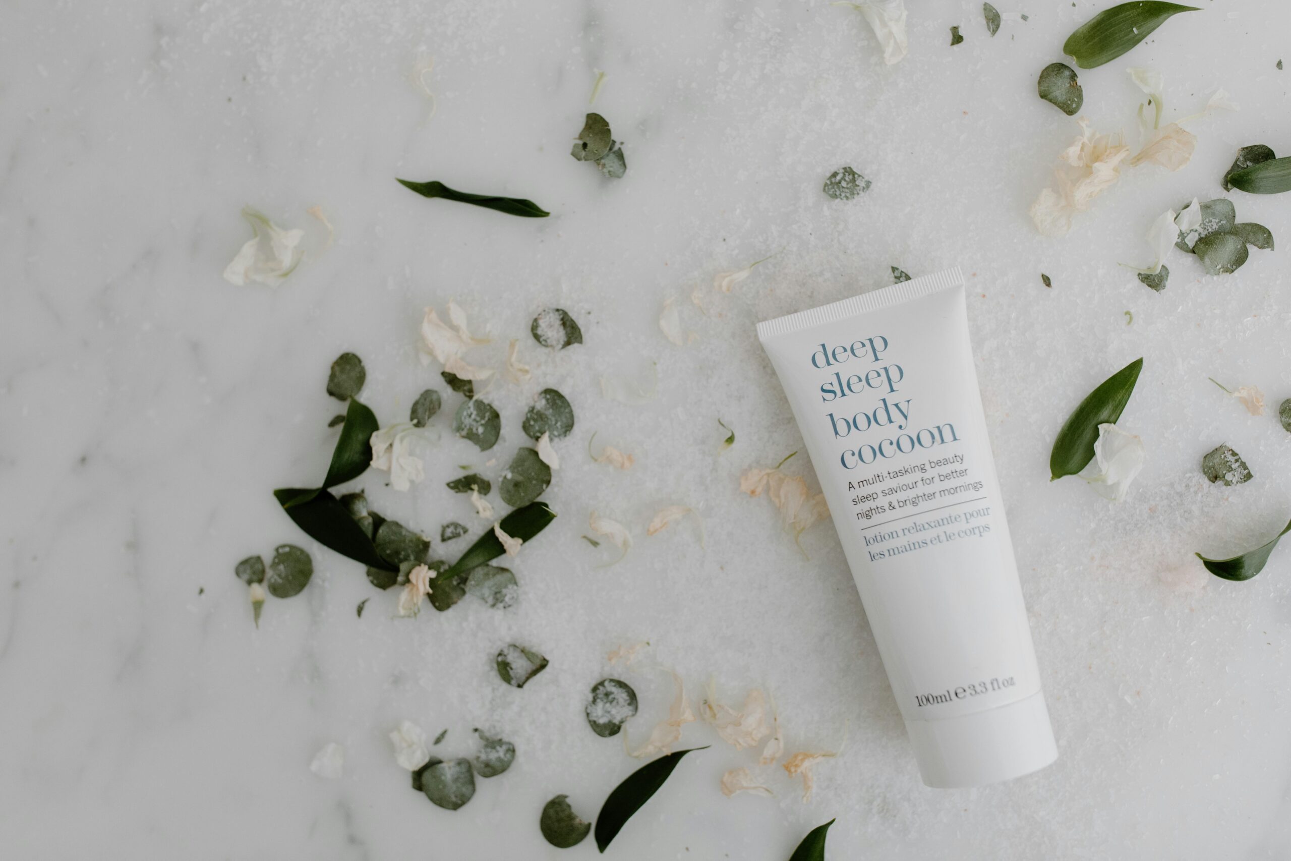 Flat lay of soothing sleep lotion surrounded by green leaves on marble, perfect for skincare themes.