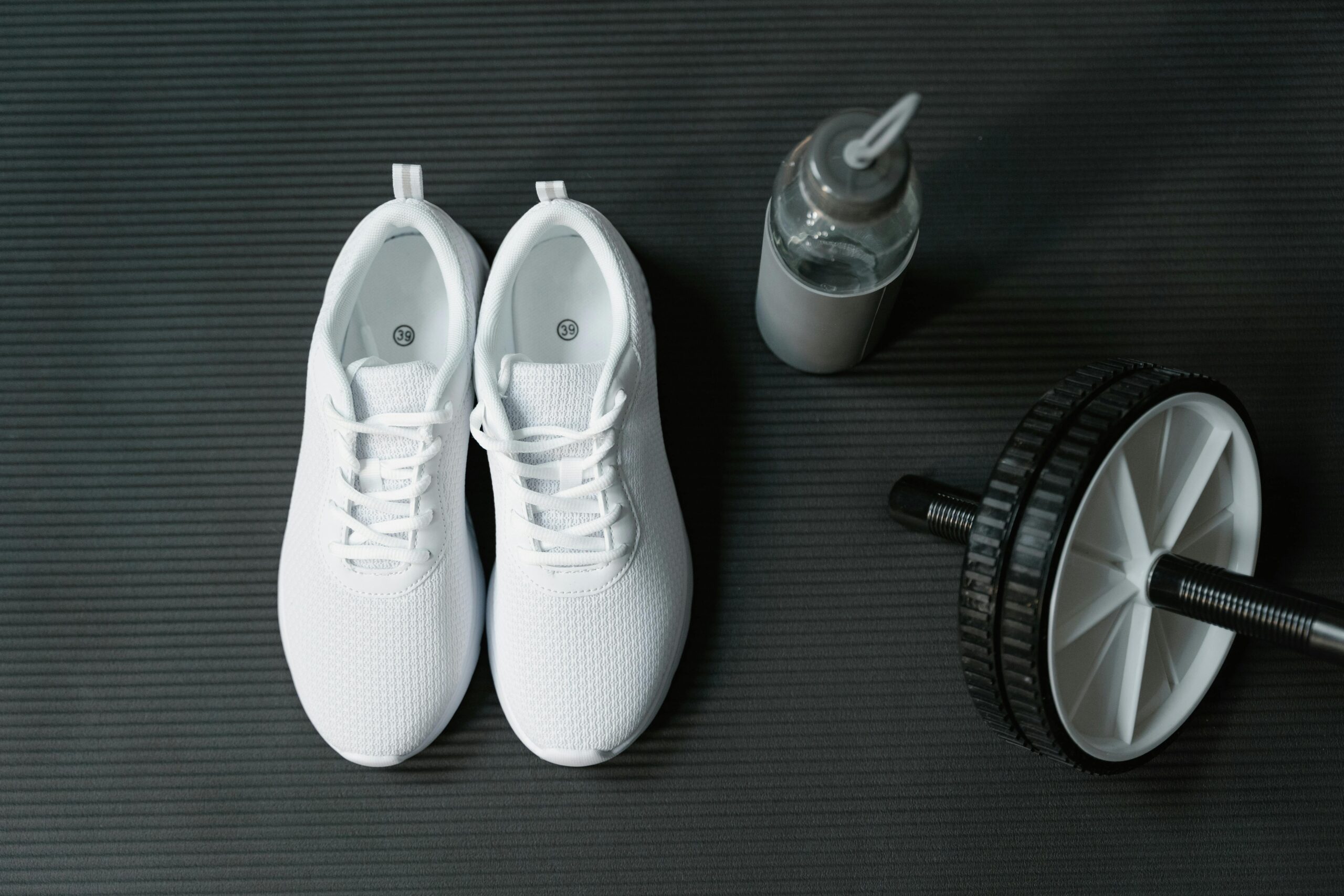 Flat lay of white sneakers, ab roller, and water bottle on black mat for fitness motivation.