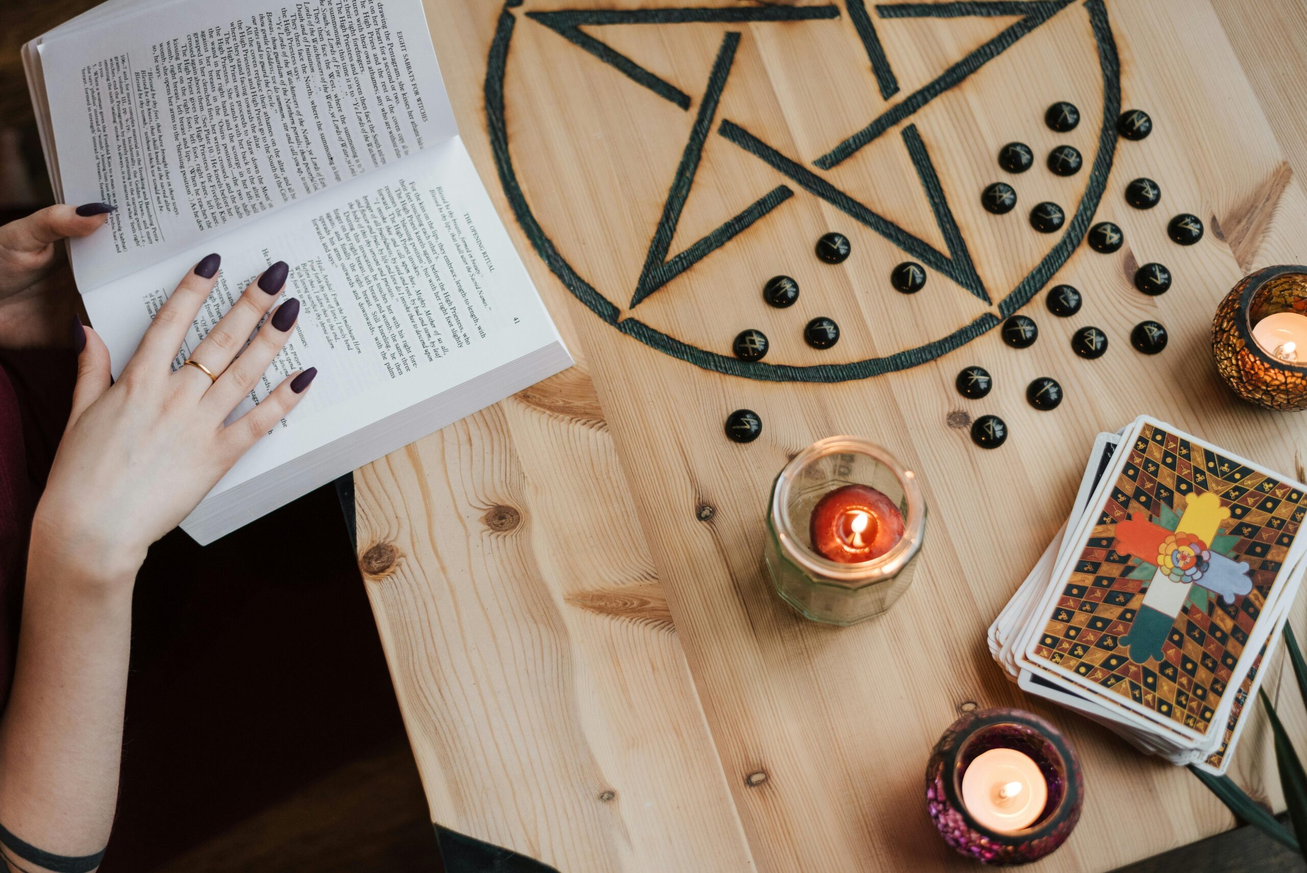 From above of crop unrecognizable fortune teller reading textbook at table with tarot cards and candles during divination process