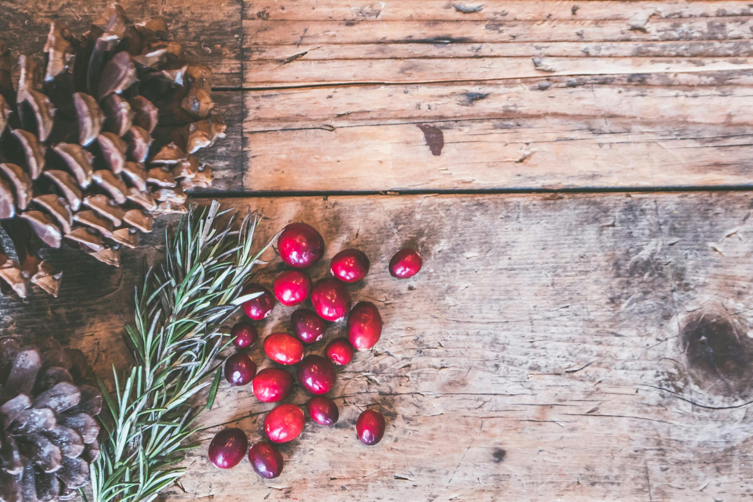 Top view of rustic holiday decor featuring pinecones, cranberries, and rosemary on a wooden surface.