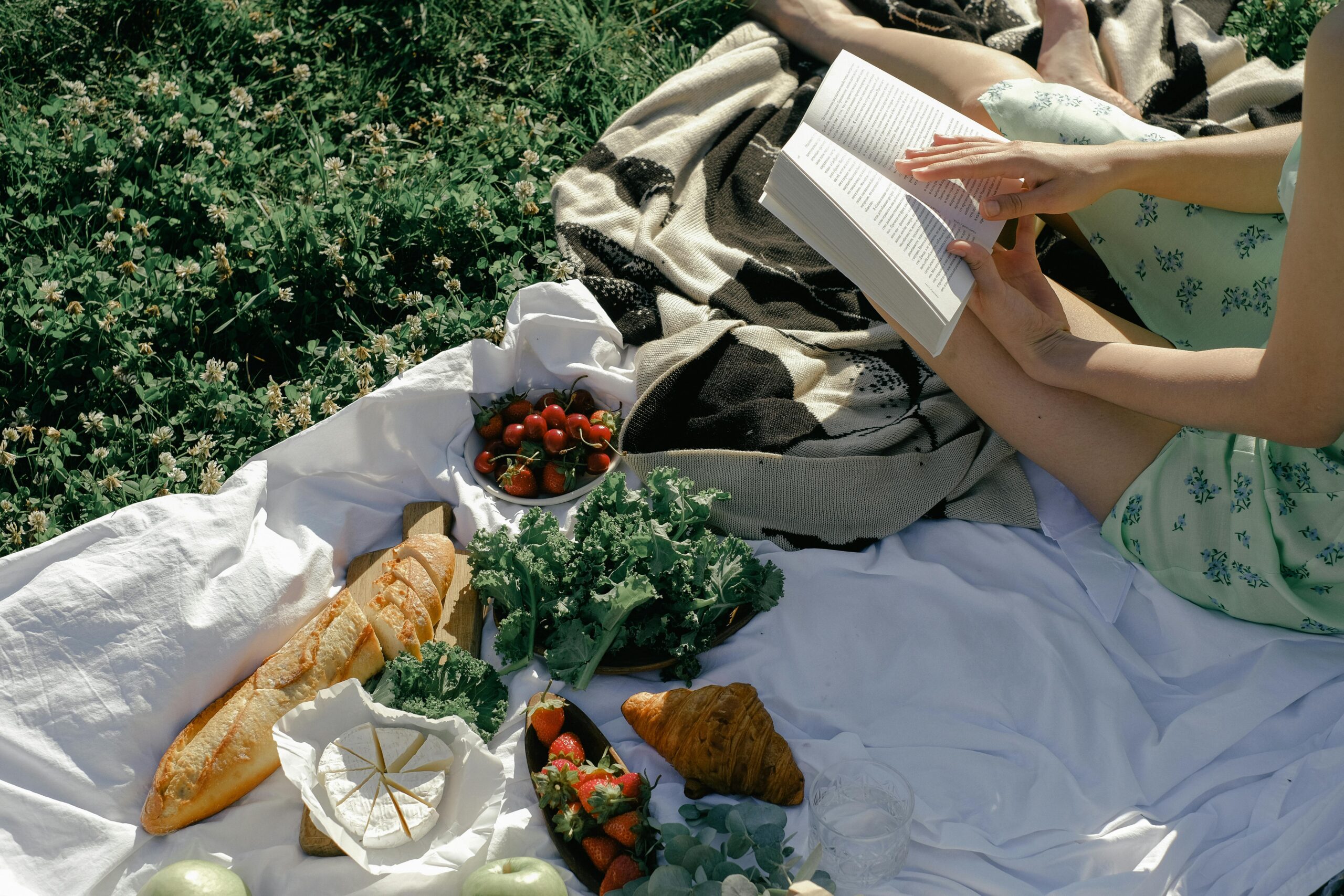 Woman enjoying a relaxing summer picnic with fresh bread, fruit, and a book on a blanket in a sunny park.