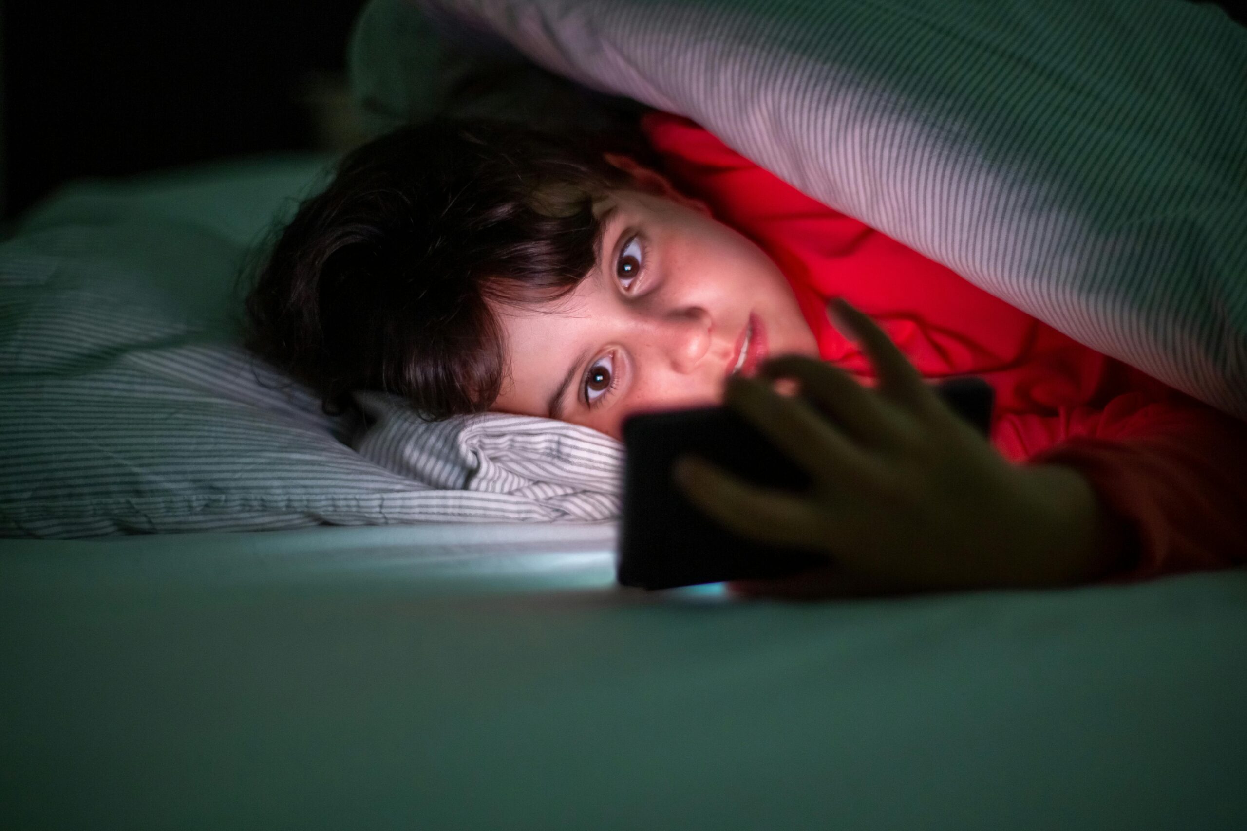 Young boy reading on smartphone under blanket at night in cozy bedroom setting.